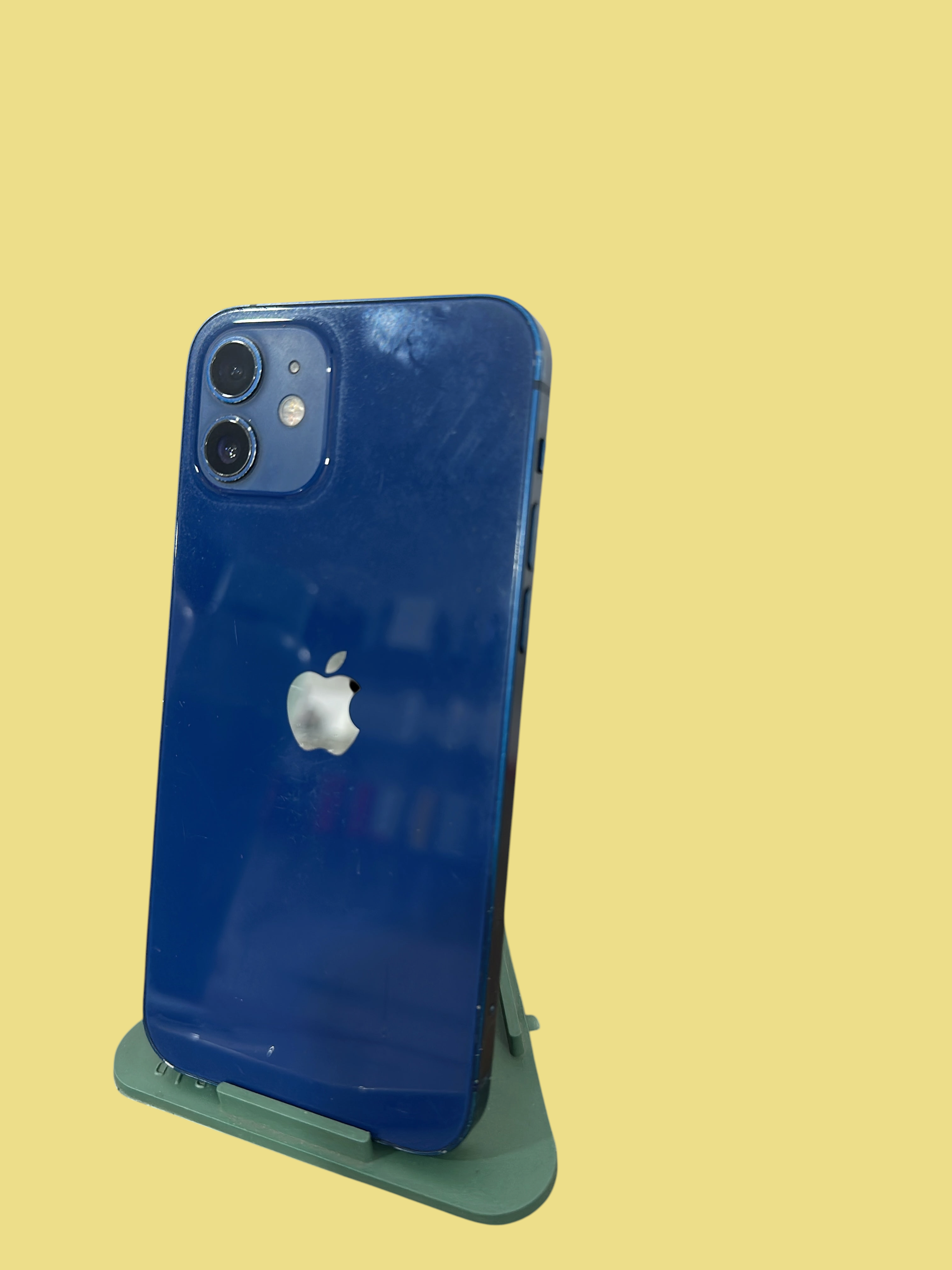 Iphoneio Official blue Apple iPhone 12 smartphone showing the dual-camera system and glass back on a green stand